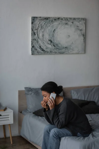 Mujer multirracial con ansiedad hablando en el teléfono inteligente en la cama en el dormitorio - foto de stock