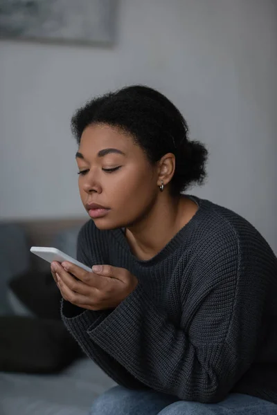 Jeune femme multiraciale utilisant un smartphone assis sur un lit flou à la maison — Photo de stock
