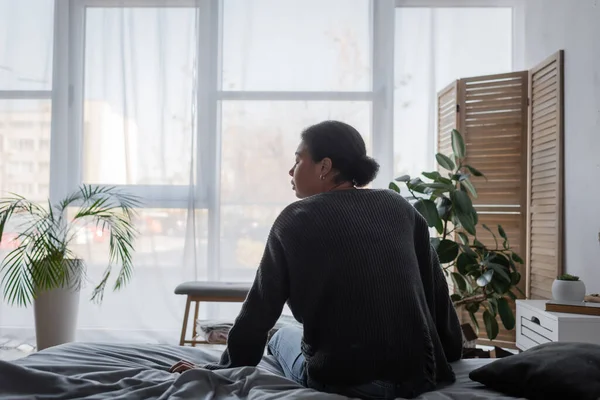 Вид сбоку грустной многорасовой женщины в вязаном свитере, сидящей дома на кровати — стоковое фото