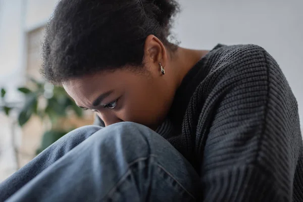 Portrait de femme multiraciale frustrée en pull et jeans assis à la maison — Photo de stock