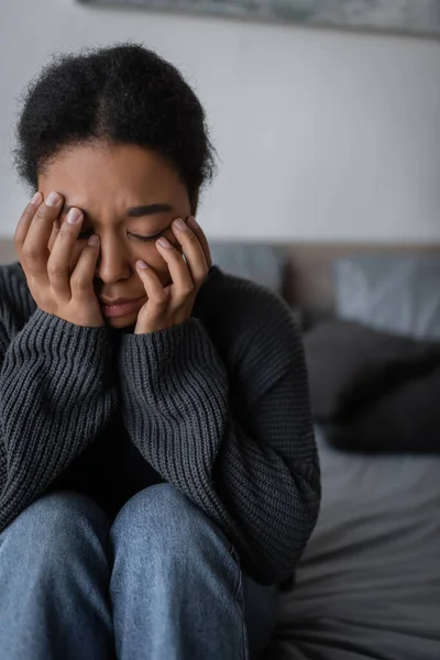 Traurige multirassische Frau im Pullover berührt Gesicht auf verschwommenem Bett zu Hause — Stockfoto