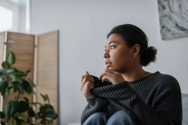 Sad multiracial woman with apathy touching knitted sweater at home — Stock Photo