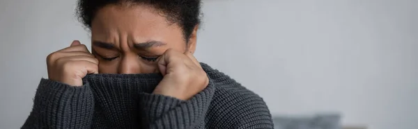 Deprimida mujer multirracial en suéter de punto llorando en casa, pancarta - foto de stock