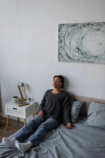 Einsame multirassische Frau mit geschlossenen Augen zu Hause auf dem Bett sitzend — Stockfoto
