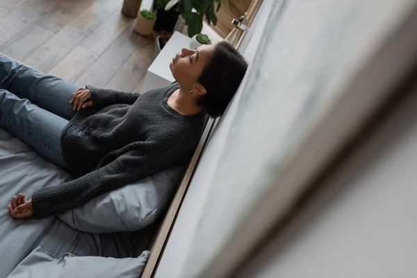 Vista ad alto angolo di donna multirazziale triste in maglione lavorato a maglia appoggiato al muro mentre seduto sul letto a casa — Foto stock