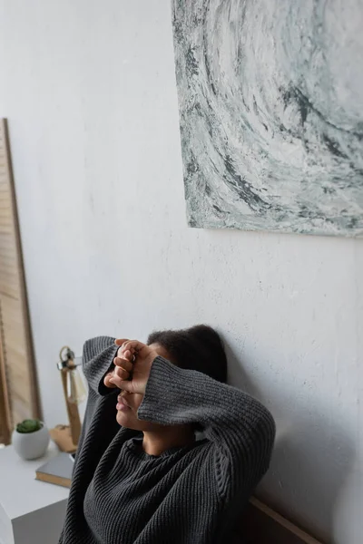 Mujer deprimida multirracial en suéter de punto que cubre los ojos en el dormitorio - foto de stock