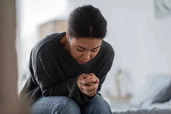 Депрессивная многорасовая женщина в трикотажном свитере смотрит на руки в спальне — стоковое фото