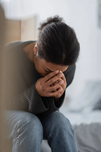 Deprimida mujer multirracial llorando mientras está sentada en la cama en casa - foto de stock