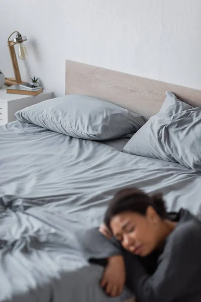 Mujer multirracial borrosa con depresión sentada cerca de la cama en casa - foto de stock