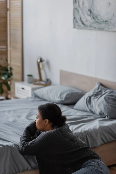 Mujer joven multirracial con problemas psicológicos mirando hacia otro lado cerca de la cama en casa - foto de stock