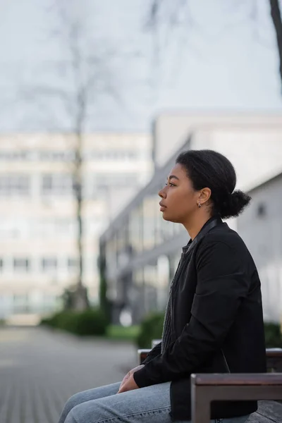 Seitenansicht einer jungen multirassischen Frau mit Depressionen, die auf einer Bank auf der städtischen Straße sitzt — Stockfoto