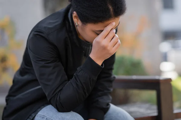 Deprimida mujer multirracial con chaqueta sentada en un banco borroso al aire libre - foto de stock