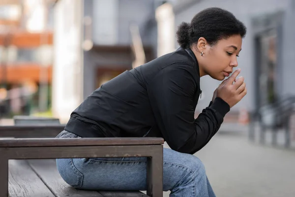 Femme multiraciale déçue en veste assise sur un banc dans une rue urbaine floue — Photo de stock