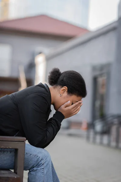 Mulher multirracial deprimida cobrindo rosto enquanto sentado no banco na rua urbana — Fotografia de Stock