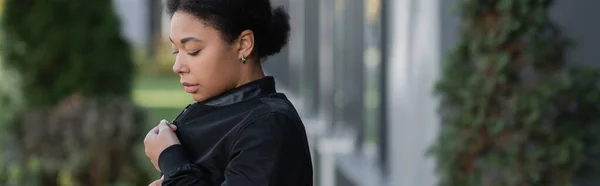Triste femme multiraciale portant une veste sur une rue urbaine floue, bannière — Photo de stock