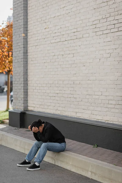 Deprimida mujer multirracial sentada en la frontera cerca del edificio al aire libre - foto de stock