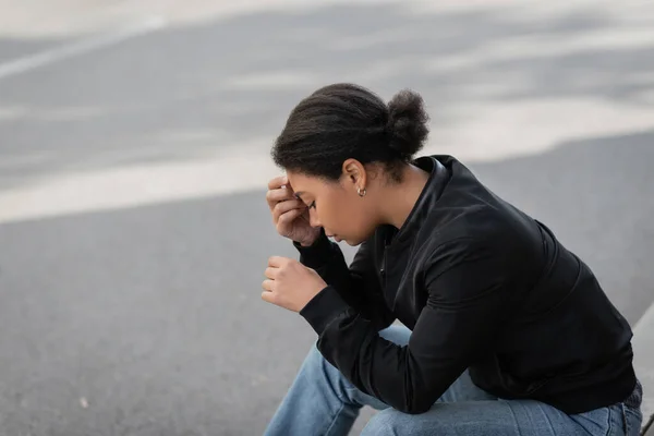 Side view of multiracial woman with apathy sitting on border on urban street — Stock Photo
