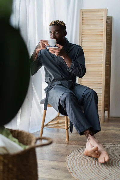 Hombre afroamericano con vitiligo en bata bebiendo café en el banco del dormitorio en casa - foto de stock