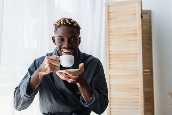 Happy african american man with vitiligo holding coffee and looking at camera at home — Stock Photo