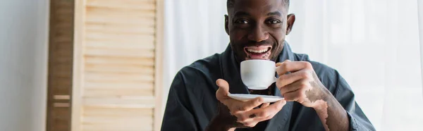Positivo hombre afroamericano con vitiligo sosteniendo la taza de café y mirando a la cámara en casa, pancarta - foto de stock