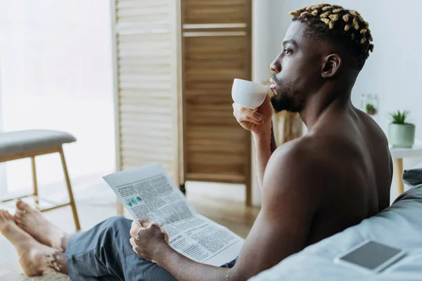 Vista lateral del hombre afroamericano con vitiligo sosteniendo café y periódico en el dormitorio - foto de stock