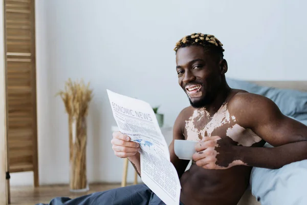 Cheerful african american man with vitiligo looking at camera and holding coffee with newspaper in bedroom — Stock Photo