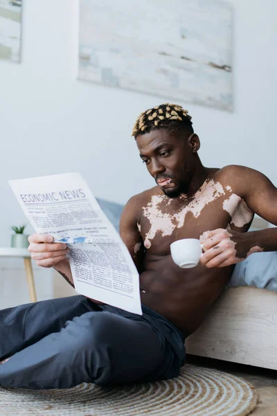 Musculoso hombre afroamericano con vitiligo sosteniendo café y leyendo el periódico en el dormitorio - foto de stock