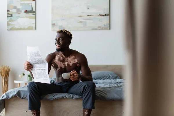 Беззаботный африканский американец с витилиго держа чашку кофе и газету, сидя на кровати дома — стоковое фото