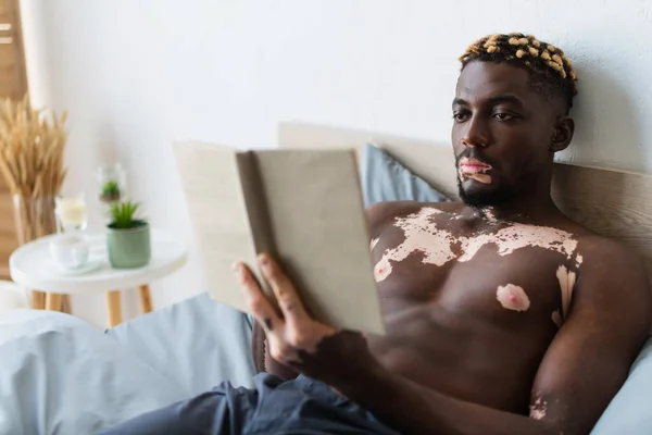 Muscular african american man with vitiligo reading blurred book on bed at home — Stock Photo