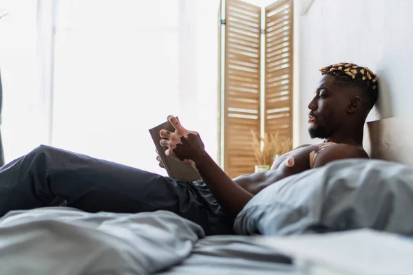Side view of shirtless african american man with vitiligo reading book in bedroom — Stock Photo