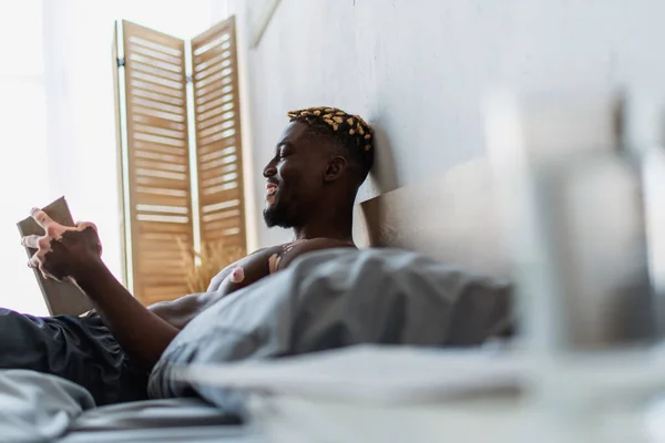 Side view of cheerful and shirtless african american man with vitiligo reading book on bed — Stock Photo