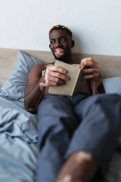 Shirtless and smiling african american man with vitiligo holding book while lying on bed at home — Stock Photo