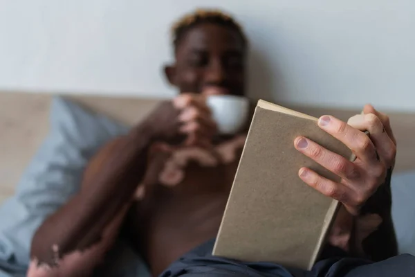 Blurred african american man with vitiligo holding cup and reading book in bed — Stock Photo
