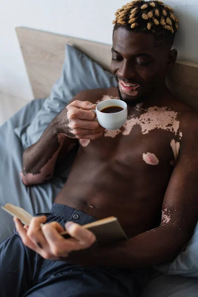 Африканский мужчина без рубашки с витилиго улыбается, держа в руках кофе и читая книгу — стоковое фото