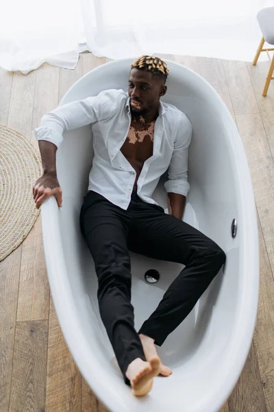Vista superior del hombre afroamericano con vitiligo en camisa y pantalones sentados en la bañera en casa - foto de stock