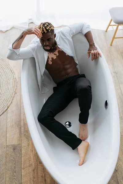 Draufsicht auf einen afrikanisch-amerikanischen Mann mit Vitiligo im Hemd, der zu Hause in der Badewanne liegt — Stockfoto