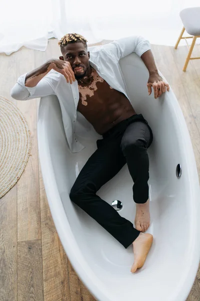 Вид сверху мускулистого африканского мужчины с витилиго в рубашке и брюках, сидящего в ванне — стоковое фото