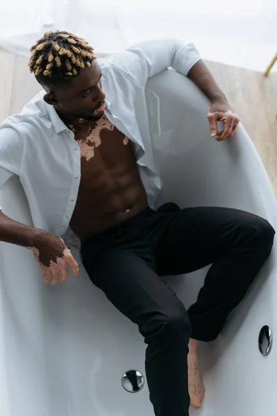 Vista aérea del hombre americano africano muscular con vitiligo sentado en la bañera en casa - foto de stock