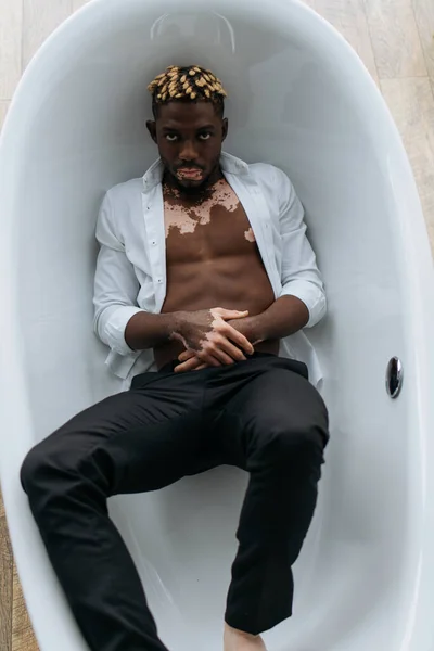 Vista superior del hombre afroamericano con vitiligo en camisa y pantalones acostados en la bañera en casa - foto de stock