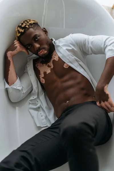 Vista superior do homem americano africano muscular com vitiligo na camisa e calças deitado na banheira em casa — Fotografia de Stock