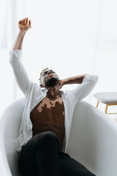 Muscular african amerian man with vitiligo in shirt sitting in bathtub at home — Stock Photo
