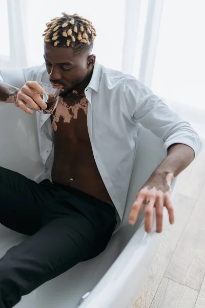 Homme afro-américain avec vitiligo en chemise blanche tenant un verre de whisky assis dans la baignoire — Photo de stock
