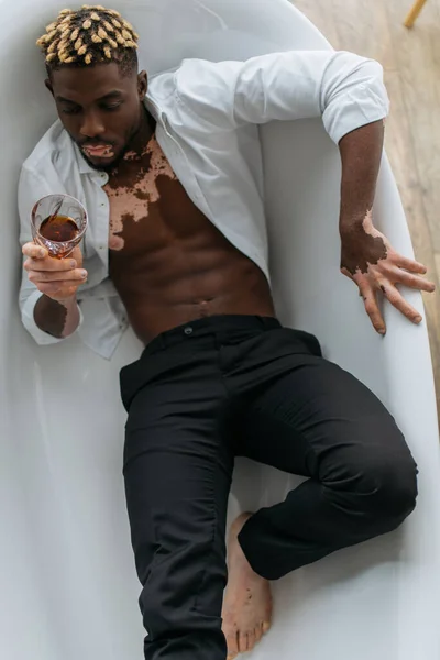 Top view of african american man with vitiligo in shirt and pants holding whiskey in bathtub — Stock Photo