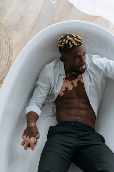 Vue du dessus de l'homme afro-américain avec vitiligo en chemise tenant du whisky tout en étant assis dans la baignoire — Photo de stock