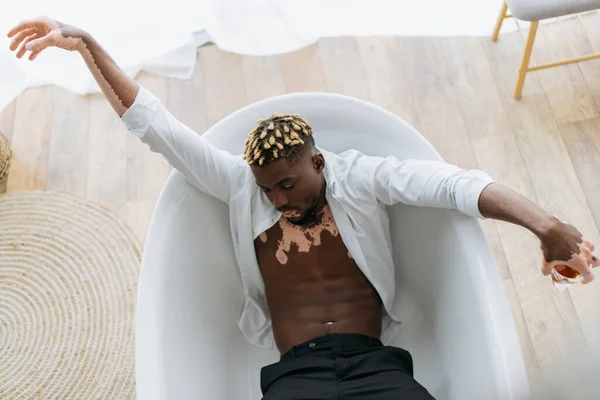 Над головою дивиться на афріканського американця з вітиліго, який тримає віскі, коли лежить у ванні. — стокове фото