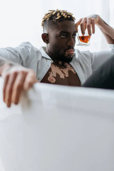 African american man with vitiligo in white shirt holding whiskey while sitting in bathtub — Stock Photo