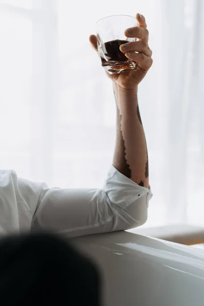 Обрезанный вид на африканского американца с витилиго в рубашке, держащего стакан виски в ванной — стоковое фото