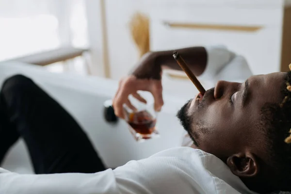 Vista lateral del hombre afroamericano con vitiligo fumando cigarro y sosteniendo whisky borroso en la bañera - foto de stock