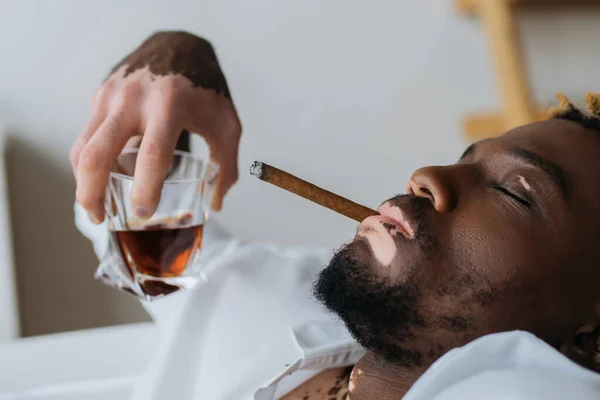Afro-américain avec vitiligo en chemise fumant du cigare et tenant du whisky dans la salle de bain — Photo de stock