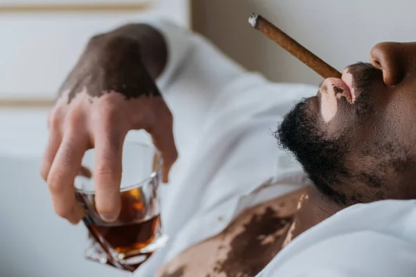 Vista recortada del hombre afroamericano con vitiligo en camisa fumando cigarro y sosteniendo whisky en el baño - foto de stock
